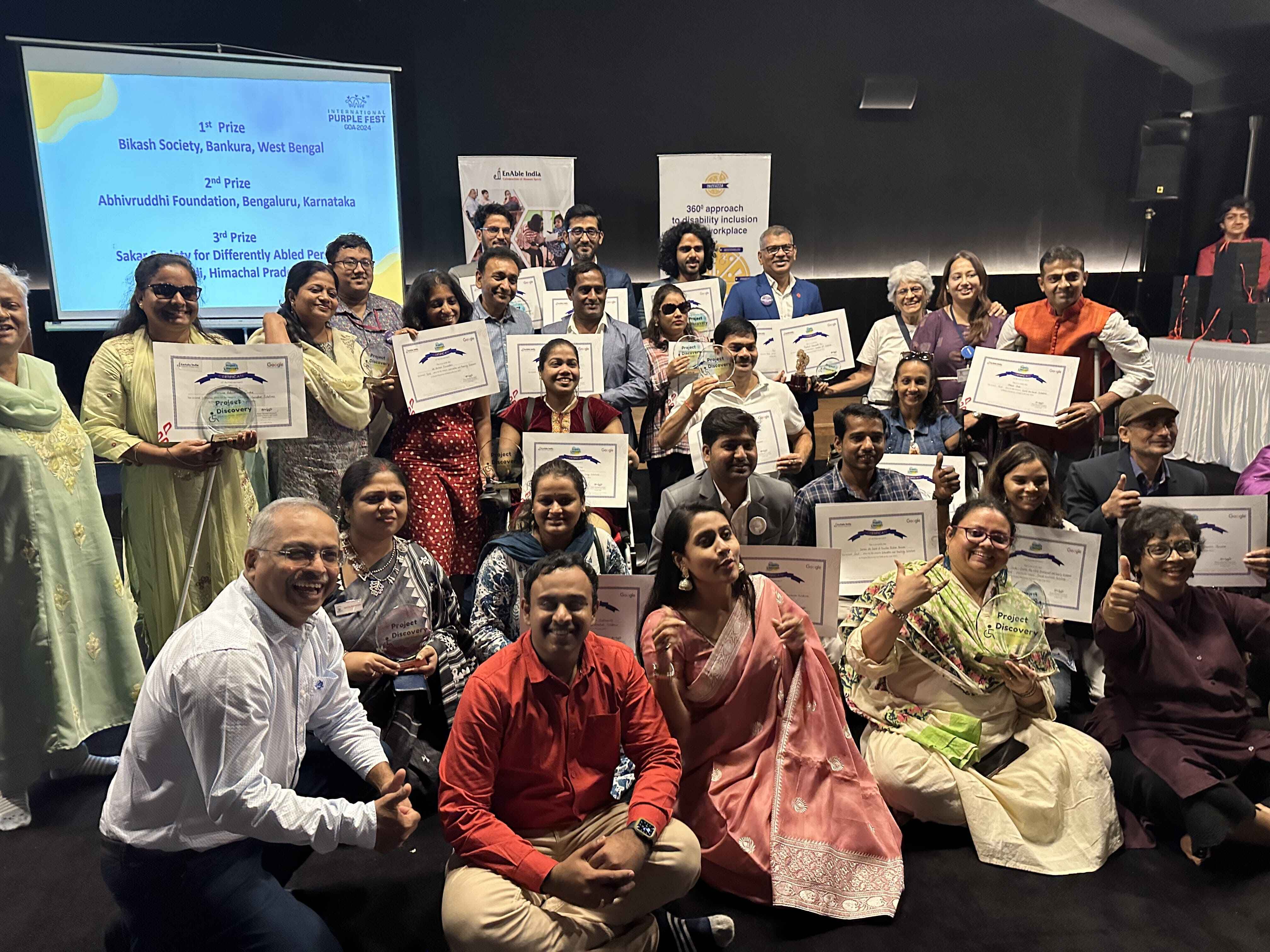 The image is a group picture from project discovery award ceremony at Purple Fest 2023.the image has been taken inside a room. The winners of Project Discovery 2023 holding their certificates. People are both standing and sitting on floor. Few winners sitting on their wheelchairs. Project Discovery team and members of senior management team an EnAble India is also present.