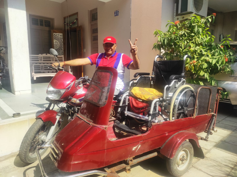 The image is of one of our innovators named Naman Patel. He is sitting and posing with his customized motorcycle and sidecar . The image is taken outside his house. The sidecar is wheelchair accessible and a folded wheelchair is visible in the sidecar.