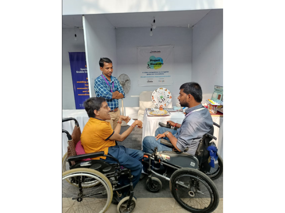 This image is taken outdoor at Purple fest 2023 and in front of project discovery solution exhibition stall. Three people are visible in the image. Two men in wheelchairs, in front of the stall, facing each other, engaged in conversation or discussion. A man standing in the stall with table in front of him with some solutions displayed. Project Discovery banner is visible on the wall behind them